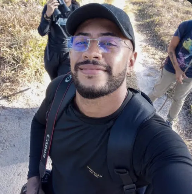 Fabrício Ferreira Jerônimo em trabalho de campo numa trilha de vegetação costeira, com câmera fotográfica a tiracolo e mochila; ao fundo, duas pessoas da equipe, uma delas observando com binóculos.